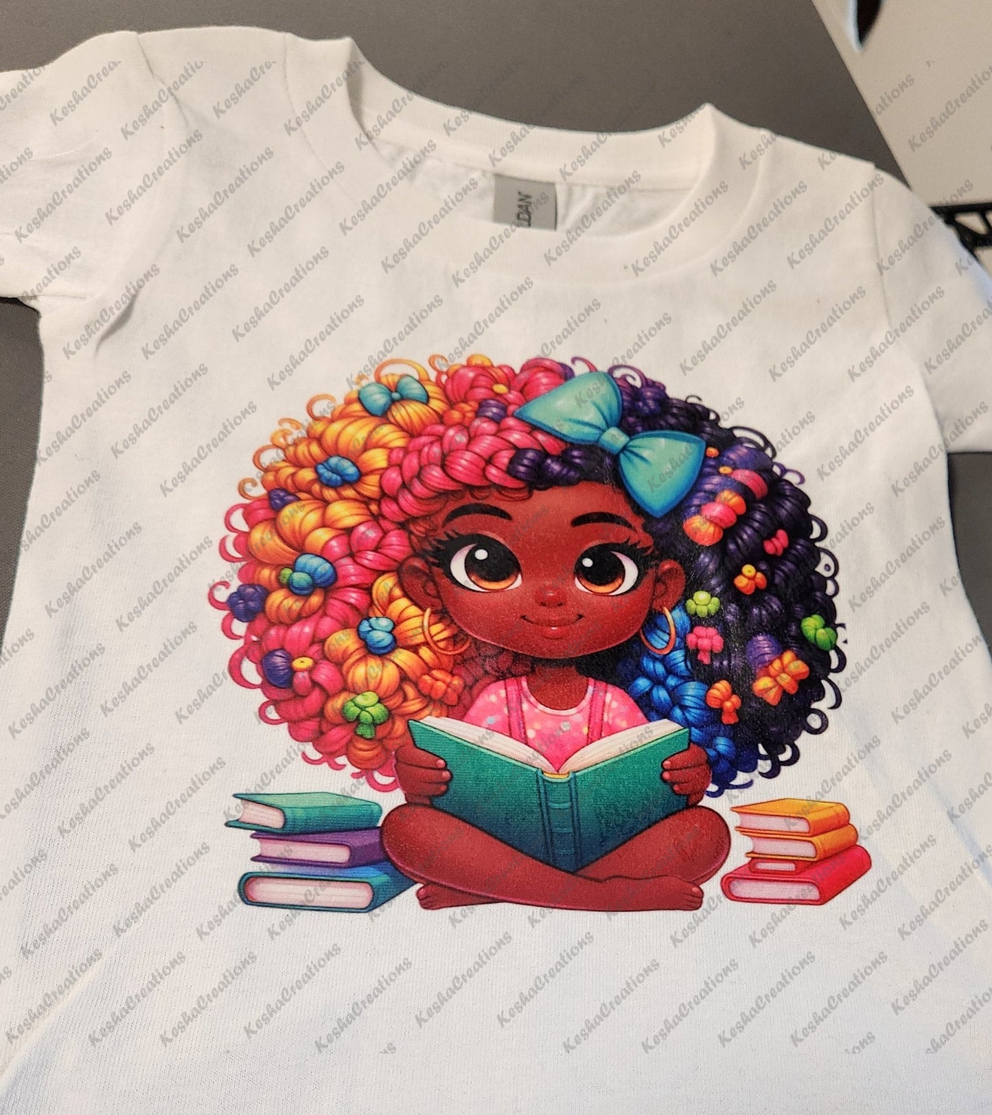 Illustration of a cute African American girl sitting cross-legged while reading a book. She has dark skin, curly rainbow hair with a blue bow, and wears a pink shirt. Several books are scattered around her as she smiles and reads.