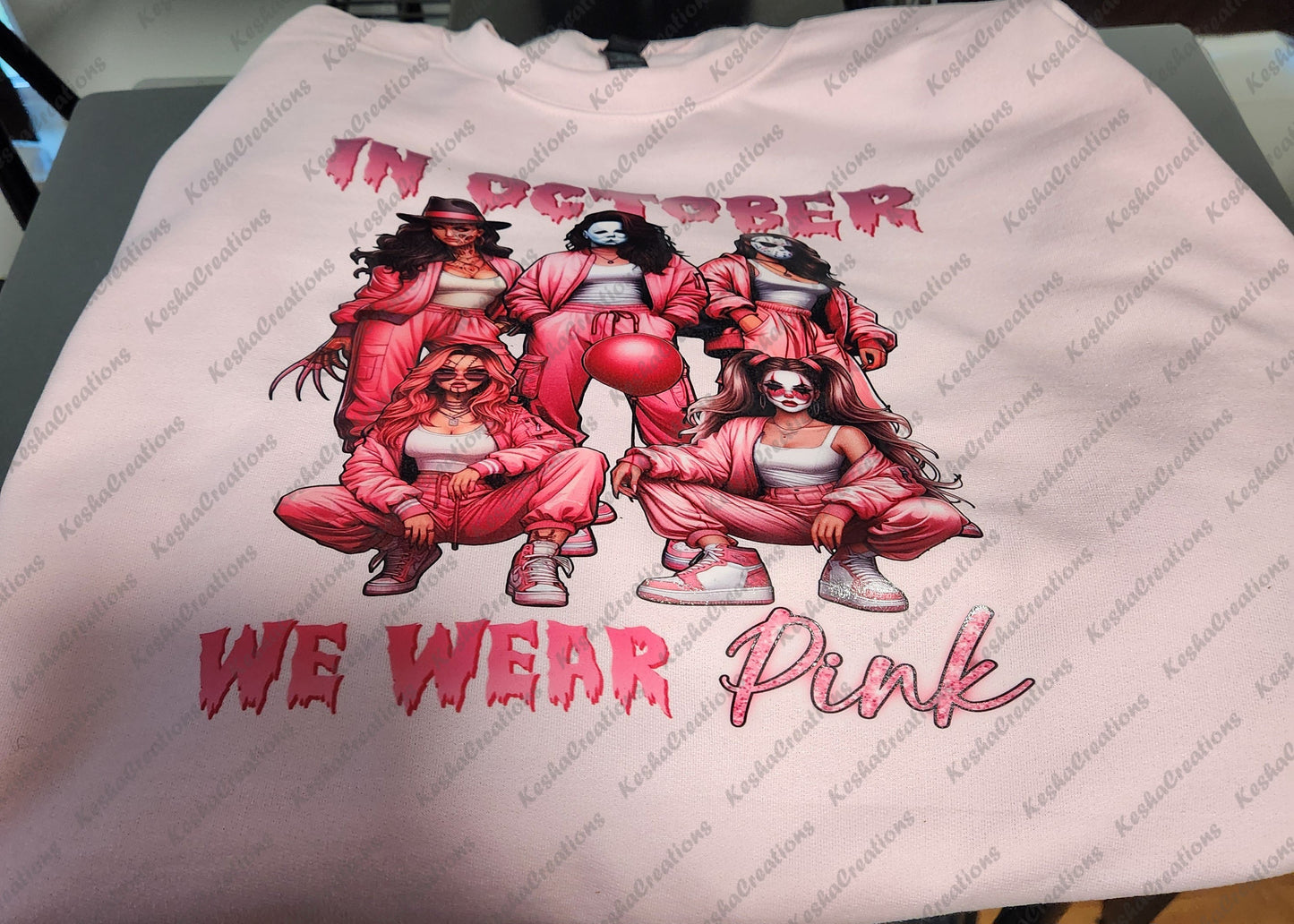 Pink sweatshirt displaying a DTF transfer design of five women dressed as horror icons, wearing pink outfits, with the slogan 'In October We Wear Pink' for breast cancer awareness.