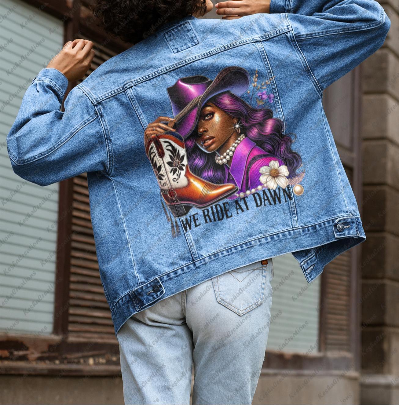 Woman posing with her back turned, wearing a denim jacket featuring a vibrant DTF transfer of a dark-skinned cowgirl with purple hair, a cowboy hat, and boots. Text below design reads "We Ride At Dawn." Showcased by KeshaCreations.