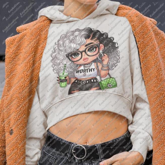 Street-style mockup featuring a woman in a cropped beige hoodie displaying a KeshaCreations DTF transfer design of a confident cartoon girl with curly black and white hair, “WORTHY” crop top, green drink, and handbag. Urban chic presentation for wearable empowerment.
